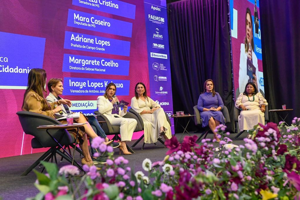 Vista lateral do palco onde cinco mulheres estão sentadas em poltronas cinzas participando de um debate. Elas vestem roupas elegantes em tons de bege, branco, azul e trajes típicos indígenas. No fundo, um painel digital azul exibe o nome das palestrantes, incluindo Adriane Lopes (Prefeita de Campo Grande) e Inaye Lopes Kaiowá. Na lateral do painel, logos de apoiadores como Sebrae, Banco do Brasil e Governo do Estado. Em primeiro plano, flores roxas e brancas decoram a borda do palco, criando um efeito levemente desfocado.