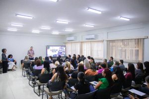 A imagem registra um momento de formação profissional em uma sala de aula bem iluminada, onde cerca de 40 pessoas, majoritariamente mulheres, estão sentadas em cadeiras com pranchetas, voltadas para um telão que exibe o título "Letramento Racial". Na frente da sala, um palestrante de camisa lilás conduz a apresentação, acompanhado por duas mulheres que o apoiam, compondo um ambiente de aprendizado e debate.