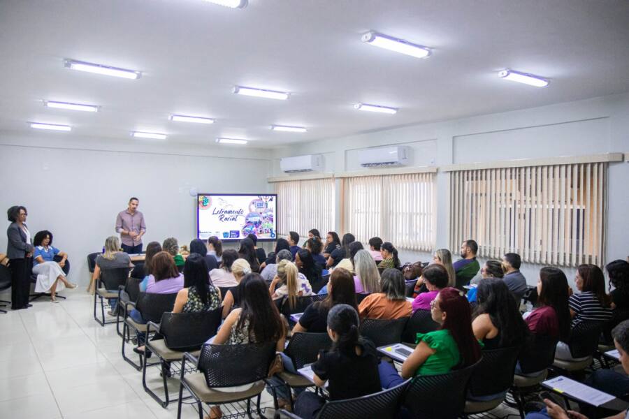 A imagem registra um momento de formação profissional em uma sala de aula bem iluminada, onde cerca de 40 pessoas, majoritariamente mulheres, estão sentadas em cadeiras com pranchetas, voltadas para um telão que exibe o título "Letramento Racial". Na frente da sala, um palestrante de camisa lilás conduz a apresentação, acompanhado por duas mulheres que o apoiam, compondo um ambiente de aprendizado e debate.