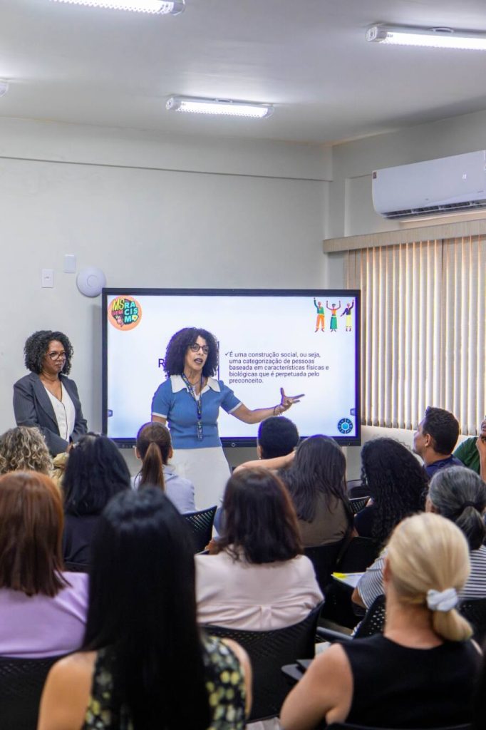 Foto de uma sala de aula com várias pessoas de costas, assistindo a uma palestra. Na frente, uma mulher negra com cabelos crespos volumosos, vestindo uma blusa azul e calça branca, fala gesticulando para uma tela de TV grande. Na tela, lê-se o trecho: "É uma construção social, ou seja, uma categorização de pessoas baseada em características físicas e biológicas que é perpetuada pelo preconceito". Ao lado dela, outra mulher negra de blazer escuro observa.