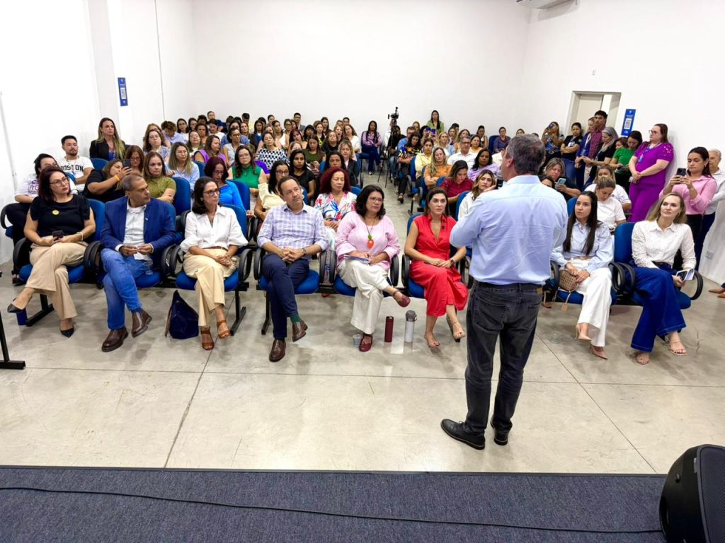 otografia em plano aberto de um auditório. Em primeiro plano, vemos as costas de um homem que fala para a plateia; ele veste camisa azul clara e calça jeans escura. Sentadas na primeira fileira, de frente para ele, estão sete pessoas (cinco mulheres e dois homens). Entre elas, destaca-se uma mulher de blusa rosa e colar de círculo vermelho ao centro, e uma mulher de vestido vermelho vibrante à direita. O restante do auditório está repleto de mulheres sentadas em cadeiras azuis, prestando atenção à fala. As paredes são brancas e o chão é de cimento queimado claro.