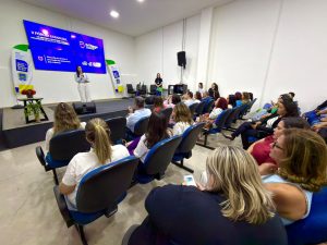 Foto tirada da lateral posterior do auditório, mostrando a plateia de costas e de perfil. As pessoas ocupam fileiras de cadeiras azuis que levam em direção ao palco no fundo. No palco, uma mulher realiza uma apresentação ao lado de um grande telão azul que exibe o título "II Fórum Estadual". É possível ver equipamentos de filmagem, como um tripé com câmera, posicionado entre os assentos. O ambiente é bem iluminado e amplo.