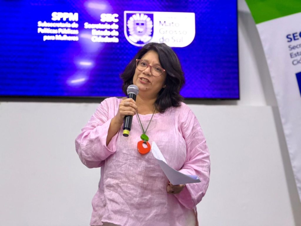 Retrato em plano médio de uma mulher de pele clara e cabelos castanhos curtos. Ela usa óculos de grau, uma blusa de linho rosa claro e um colar com um pingente circular grande e vermelho. Ela segura um microfone na mão direita e algumas folhas de papel na mão esquerda, com uma expressão de fala. Ao fundo, há um painel digital azul com logotipos brancos, incluindo a sigla "SPPM" (Subsecretaria de Políticas Públicas para Mulheres) e o brasão do Estado de Mato Grosso do Sul.