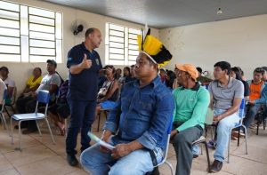Fotografia em um ambiente interno simples com teto de metal e janelas altas. No centro, um homem indígena de perfil veste uma camisa de jeans azul e um cocar de penas amarelas e pretas. Atrás dele, à esquerda, um homem de camiseta polo azul marinho gesticula com o polegar para cima enquanto fala com o grupo. Diversas pessoas, homens e mulheres de origem indígena, estão sentadas em cadeiras azuis ao fundo, observando a interação.
