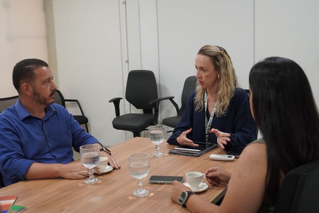 Três pessoas em uma reunião ao redor de uma mesa de madeira. No centro, uma mulher loira de blusa azul marinho fala com as mãos. À esquerda, um homem de camisa azul a escuta atentamente, e à direita, uma mulher aparece de costas. Sobre a mesa, há copos de água, xícaras de café e um tablet.