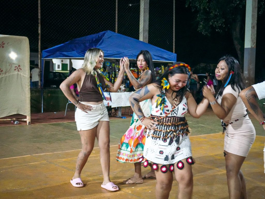 Fotografia noturna mostrando quatro mulheres dançando em uma quadra iluminada. Elas sorriem e celebram. Algumas usam roupas casuais e outras vestem trajes com grafismos e adereços típicos, como sementes e penas. Uma das mulheres tem pinturas corporais nos braços. Ao fundo, vê-se uma tenda azul e a estrutura da quadra sob o céu escuro.