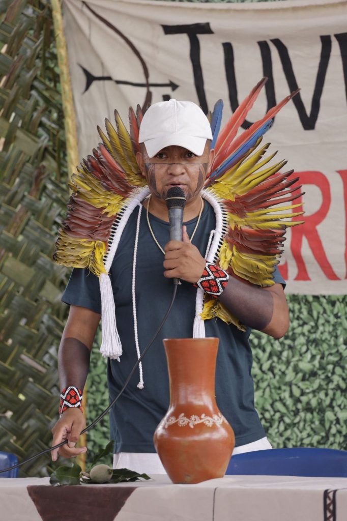 Retrato centralizado de um jovem indígena de frente para a câmera. Ele veste uma camiseta de cor vinho com um logotipo circular no peito esquerdo que menciona "Juventude Terena - Cachoeirinha". Ele usa um grande cocar de penas em tons de amarelo, laranja e marrom, com detalhes em cordões brancos que caem sobre o peito. Ao fundo, há um painel decorativo e um céu azul.