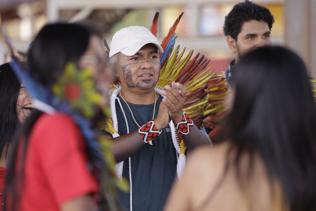 Foto em close de um homem indígena sorridente durante uma celebração. Ele usa um boné branco, pintura facial preta na região da mandíbula e um imponente cocar de penas amarelas e vermelhas que desce pelos ombros. Ele está com as mãos juntas, como se batesse palmas, e usa pulseiras com grafismos indígenas em vermelho e preto. Outras pessoas aparecem desfocadas ao redor.