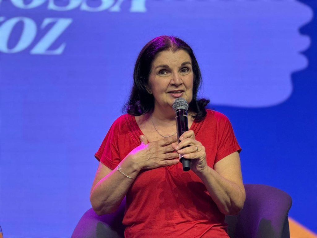 Foto em close de uma mulher de blusa vermelha e cabelos escuros, sentada em uma poltrona cinza. Ela segura o microfone próximo à boca com a mão direita e repousa a mão esquerda sobre o peito, em um gesto de emoção ou sinceridade enquanto fala.