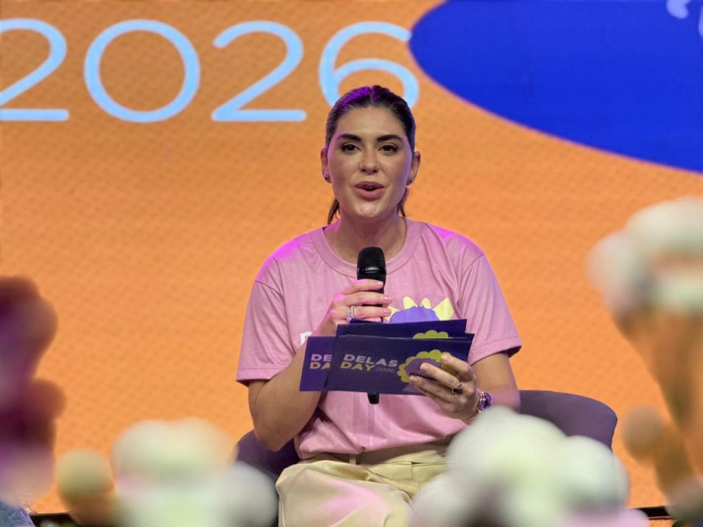 Foto em close de uma mulher branca de cabelos castanhos presos, vestindo uma camiseta rosa do evento. Ela segura um microfone com as duas mãos e olha para a frente com uma expressão comunicativa. Ao fundo, parte do telão exibe o ano "2026".