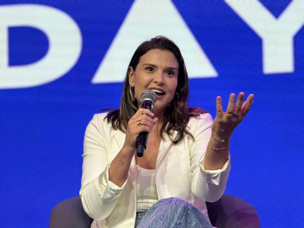 Foto em close de uma mulher de blazer branco e cabelos castanhos soltos. Ela fala ao microfone com entusiasmo, gesticulando com a mão esquerda aberta. Ela usa anéis dourados e uma pulseira delicada. O fundo é azul vibrante.
