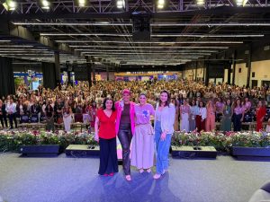 Foto em plano aberto do palco para a plateia. Quatro mulheres estão em pé no centro do palco, sorrindo para a foto. À frente delas, uma borda de flores naturais em tons de rosa e branco. O auditório está lotado com centenas de mulheres sentadas em cadeiras organizadas em fileiras. Ao fundo, brilha um letreiro que diz "Mercado Sebrae". O teto é alto, com estrutura industrial e iluminação de palco profissional.