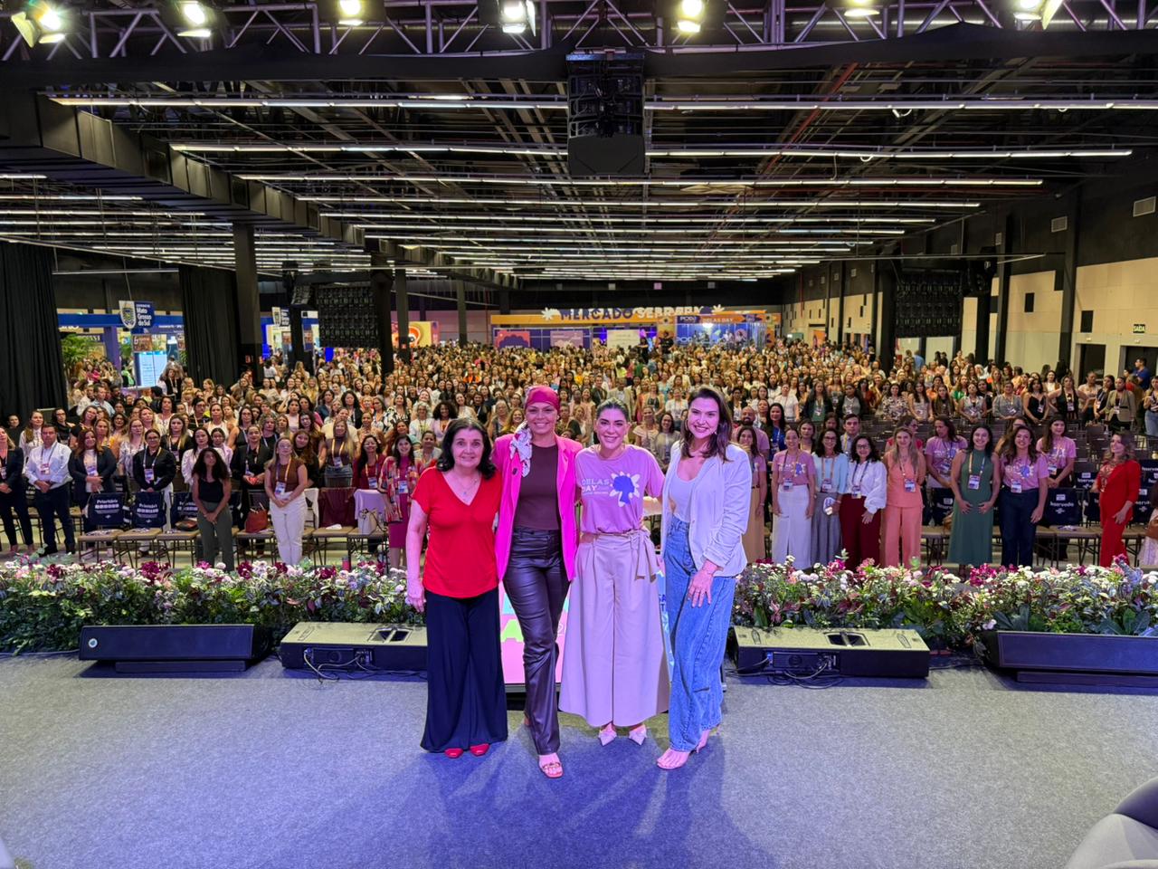 Foto em plano aberto do palco para a plateia. Quatro mulheres estão em pé no centro do palco, sorrindo para a foto. À frente delas, uma borda de flores naturais em tons de rosa e branco. O auditório está lotado com centenas de mulheres sentadas em cadeiras organizadas em fileiras. Ao fundo, brilha um letreiro que diz "Mercado Sebrae". O teto é alto, com estrutura industrial e iluminação de palco profissional.