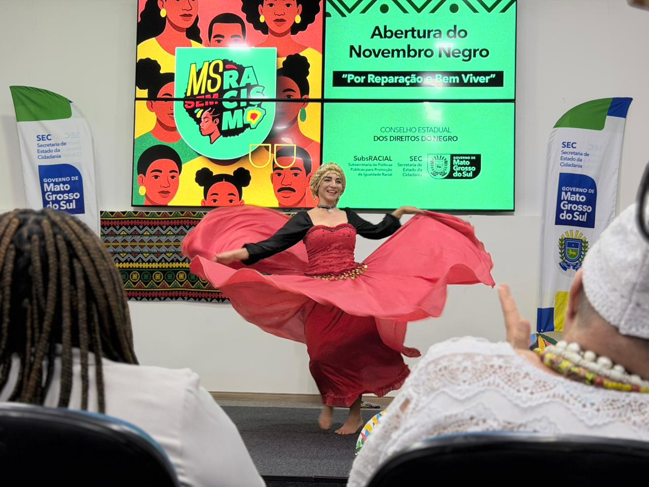 Uma dançarina no centro de um palco realiza uma performance. Ela veste uma blusa preta de mangas longas e uma saia longa e volumosa de cor rosa intenso, que ela segura pelas extremidades, criando um movimento circular. Ao fundo, uma grande tela exibe a frase "Abertura do Novembro Negro - Por Reparação e Bem Viver", ilustrada com rostos de pessoas negras em estilo artístico. Nas laterais, há bandeiras do Governo de Mato Grosso do Sul. Em primeiro plano, vemos as costas de pessoas da plateia assistindo à apresentação.