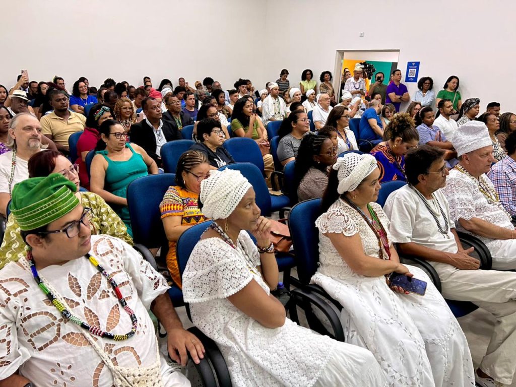 Fotografia em plano aberto mostrando o público sentado em um auditório. A plateia é diversa, composta por homens e mulheres de diferentes idades. Na primeira fila, destacam-se pessoas vestindo roupas brancas tradicionais e contas (guias) no pescoço, vestimentas típicas de religiões de matriz africana. O ambiente tem paredes brancas e cadeiras azuis, estando quase completamente lotado.