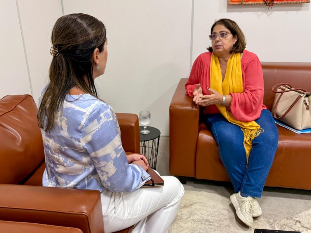 Foto de duas mulheres sentadas em poltronas de couro marrom, conversando em um ambiente interno. À direita, a Ministra Cida Gonçalves, uma mulher de pele clara e óculos, veste uma blusa coral com um lenço amarelo vibrante sobre os ombros e calça jeans. Ela gesticula com as mãos enquanto fala. À esquerda, de costas para a câmera, a secretária da Cidadania, Viviane Luiza, de cabelos castanhos presos observa, vestindo uma blusa estampada em tons de azul e branco e calça branca.