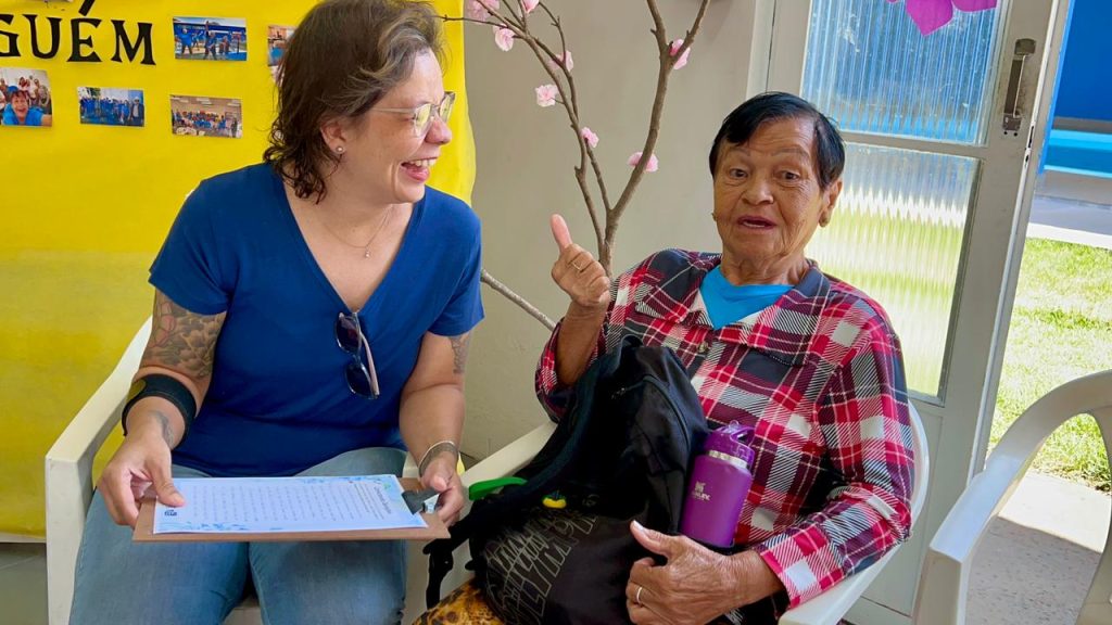 Uma mulher jovem, de óculos e camiseta azul, sorri alegremente enquanto segura uma prancheta com um documento. Ela está sentada ao lado de uma senhora idosa em uma cadeira de plástico branca. A senhora veste uma blusa xadrez vermelha e preta, sorri para a câmera e faz um sinal de "joinha" com o polegar. Ao fundo, uma parede amarela com fotos murais e um pequeno galho decorativo com flores rosas.
