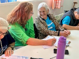 Close de duas mulheres sentadas à mesa. À esquerda, uma mulher com cabelos ruivos ondulados e camisa verde limão escreve em uma folha de papel sobre uma prancheta. À direita, uma senhora idosa com cabelos brancos e casaco de estampa de oncinha olha para a mulher ruiva com um sorriso carinhoso e atento. Sobre a mesa, há uma garrafa de água roxa e outros papéis.