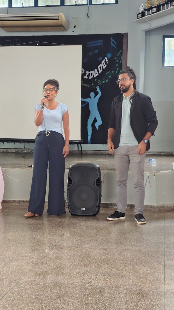 Em plano médio, um homem e uma mulher estão de pé no centro do salão, à frente do palco. A mulher, à esquerda, tem pele negra, usa óculos, blusa azul-clara e calça pantalona escura; ela segura um microfone e fala. O homem, à direita, tem pele morena, usa óculos, barba, cabelo preso, camisa clara e blazer escuro. Entre eles, no chão, há uma caixa de som preta.
