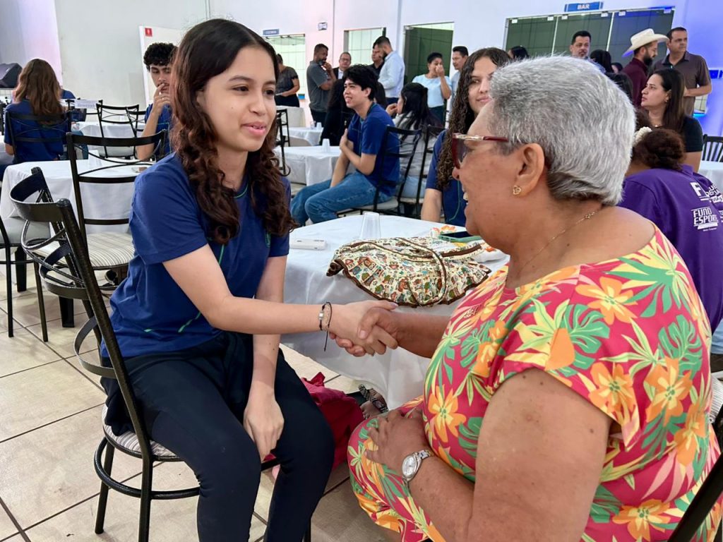 Uma jovem e uma senhora idosa estão sentadas de frente uma para a outra, apertando as mãos e sorrindo. A jovem veste uma camiseta azul e a senhora usa uma blusa com estampa floral colorida e óculos. O gesto simboliza o encontro e o respeito entre as gerações. Ao fundo, é possível ver outras mesas e participantes do evento em um ambiente amplo e bem iluminado.