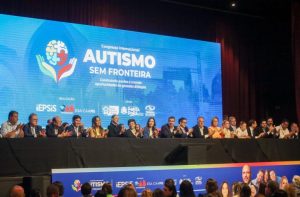 Uma perspectiva mais ampla do palco principal. Uma fileira extensa de autoridades (cerca de 20 pessoas) está de pé ou sentada, aplaudindo. O telão ao fundo reforça o tema: "Autismo Sem Fronteira - Construindo pontes e criando oportunidades de grandes diálogos". Na base do palco, há um banner com fotos de palestrantes e logotipos de apoiadores.