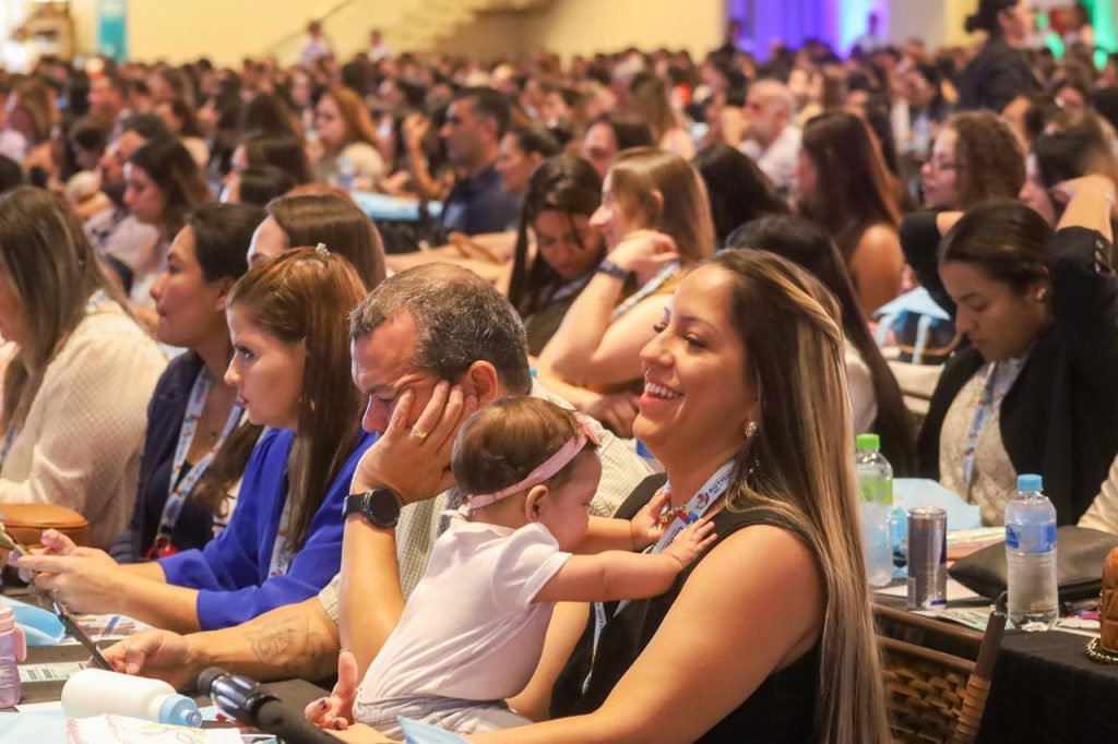 Close-up de uma seção da plateia. Em destaque, uma mulher sorridente segura um bebê no colo enquanto acompanha a palestra. Ao redor, outros participantes observam atentamente, alguns fazendo anotações ou usando o celular. A atmosfera é de acolhimento e interesse.