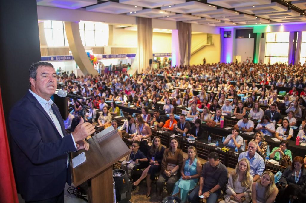 Um homem de terno escuro e camisa clara discursa em um púlpito de madeira. Ele gesticula com a mão esquerda enquanto fala ao microfone. Ao fundo, a imagem mostra a profundidade do salão repleto de pessoas sentadas, criando um contraste entre o palestrante em primeiro plano e a grande audiência.