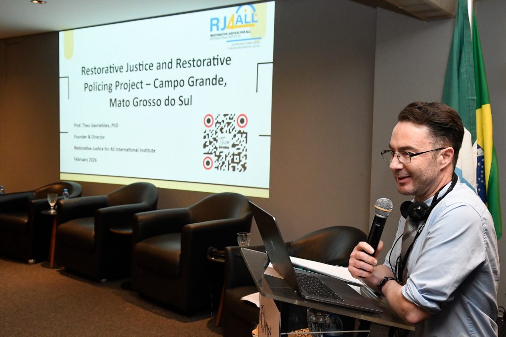 O Prof. Theo Gavrielides, PhD, Fundador e Diretor do RJ4ALL International Institute, é visto palestrando sobre o "Restorative Justice and Restorative Policing Project – Campo Grande, Mato Grosso do Sul". Ele está em pé em um pódio, usando um microfone, com um laptop aberto à sua frente e a projeção da apresentação ao fundo.