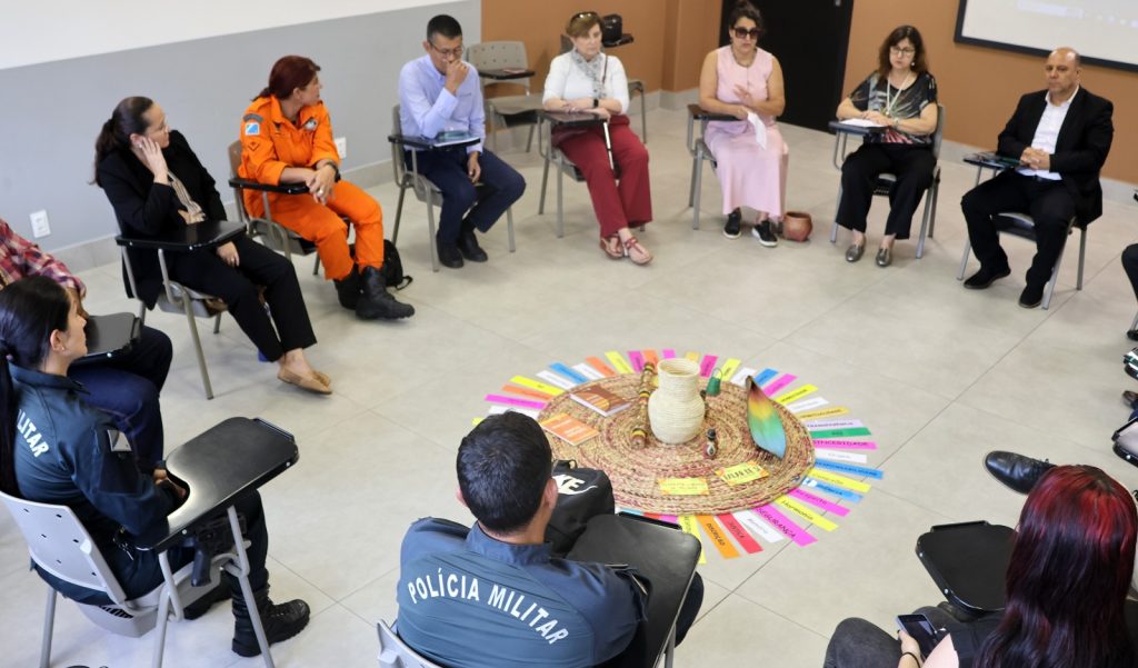 Close-up de um tapete circular de palha trançada disposto no chão. Sobre ele, repousam objetos da cultura indígena: um maracá de cabaça à esquerda, um pequeno vaso de cerâmica marrom com detalhes brancos ao centro, e uma peça artesanal adornada com penas escuras e amarelas ao fundo, dentro de um cesto raso. O fundo é o piso cinza claro da sala.