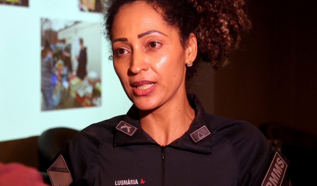 Retrato em close de uma policial militar (identificada pelo nome "Lusmária" e a insígnia "PMMS" no uniforme) falando. Ela tem pele parda, cabelos cacheados presos em um coque alto e usa brincos pequenos. A iluminação é focada em seu rosto, destacando sua expressão durante a fala. Ao fundo, uma imagem projetada aparece levemente desfocada.