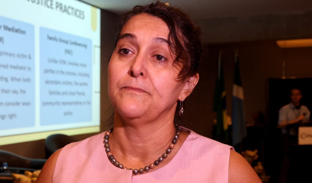 Fotografia em plano médio de uma mulher de pele clara e cabelos escuros presos, levemente ondulados na frente. Ela veste uma blusa rosa claro e um colar de pérolas em tons de cinza e rosa. Ela olha para o lado com uma expressão séria e atenta. Ao fundo, à esquerda, há uma tela de projeção com o título "JUSTICE PRACTICES" e textos explicativos sobre Mediação e Conferência de Grupos Familiares.