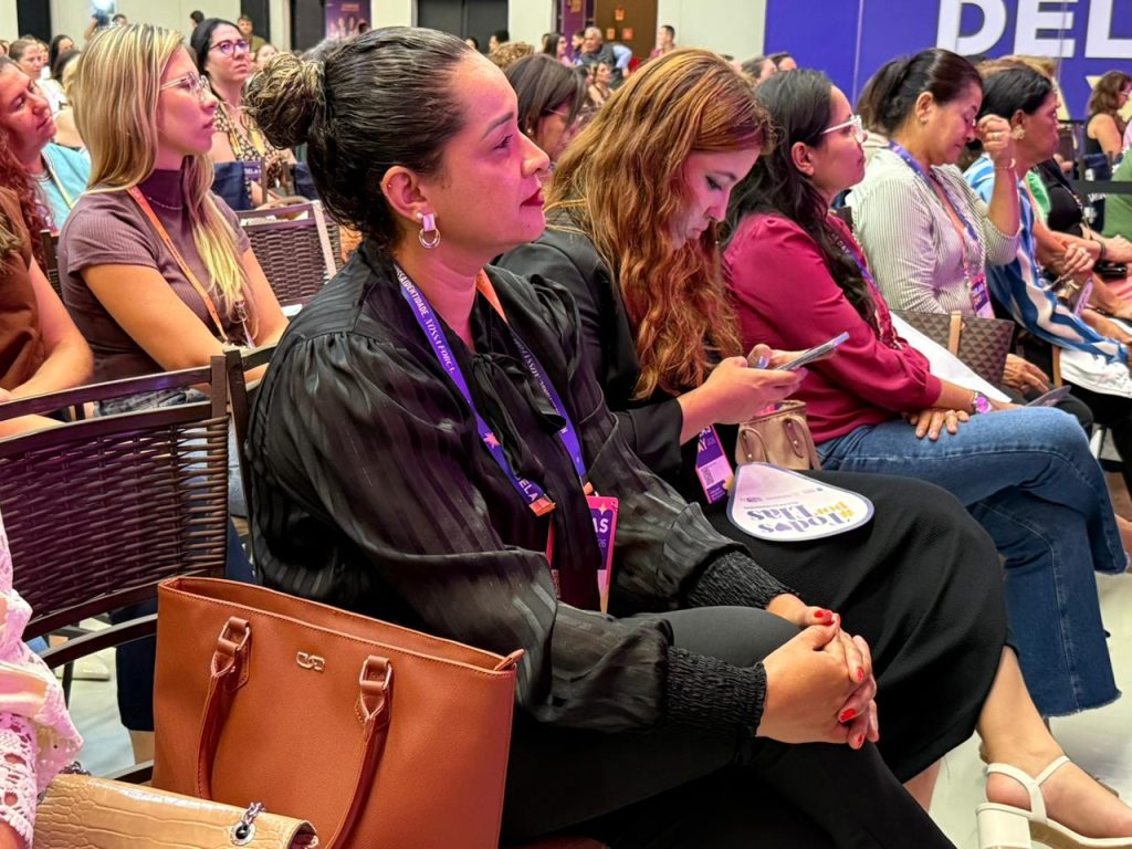 Foto em ângulo lateral focando em uma mulher na plateia. Ela veste uma blusa preta acetinada, tem os cabelos presos em um coque e usa um crachá do evento. Ela observa o palco com atenção, com as mãos entrelaçadas no colo. Ao lado, outras mulheres aparecem focadas em seus celulares ou assistindo à palestra.