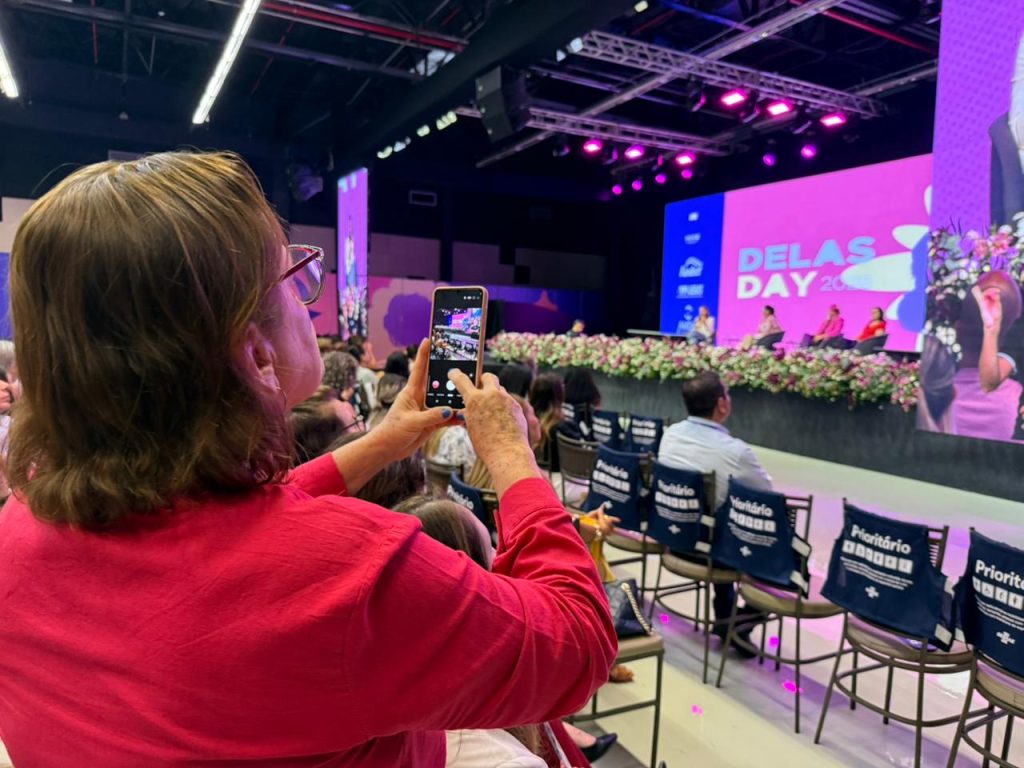 Foto em close por trás de uma mulher de blusa rosa pink que levanta o celular para fotografar ou filmar o palco. A tela do celular mostra o palco iluminado e as palestrantes. As cadeiras ao redor possuem capas azuis com a palavra "Prioritário" escrita em branco.