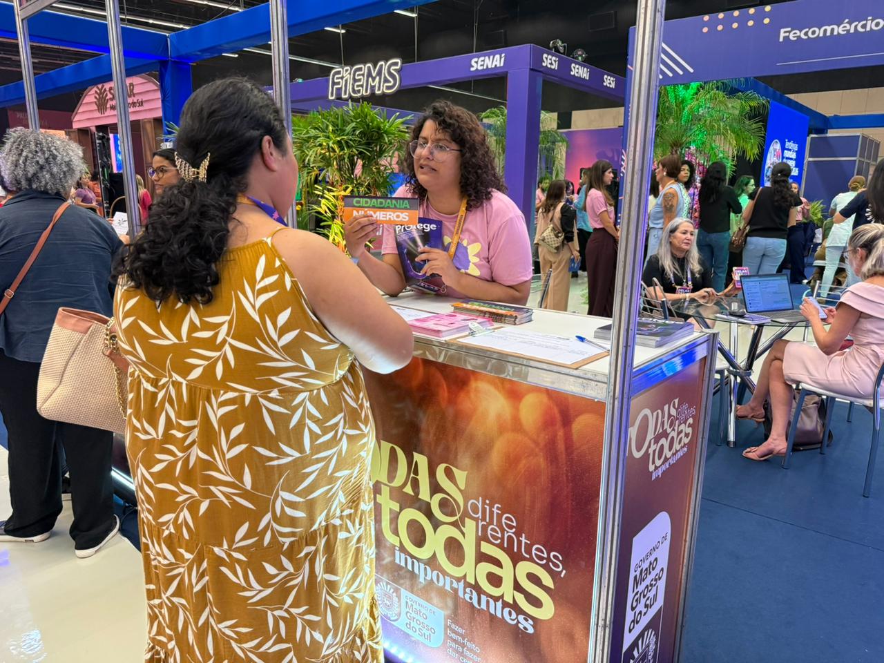 Em um ambiente de feira ou exposição, uma atendente de camiseta rosa e óculos conversa com uma visitante de costas, que usa um vestido amarelo com estampa de folhagens. Elas estão em um balcão com a marca "Delas: diferentes, todas importantes". A atendente segura materiais informativos. Ao fundo, veem-se estandes de instituições como FIEMS e SENAI.