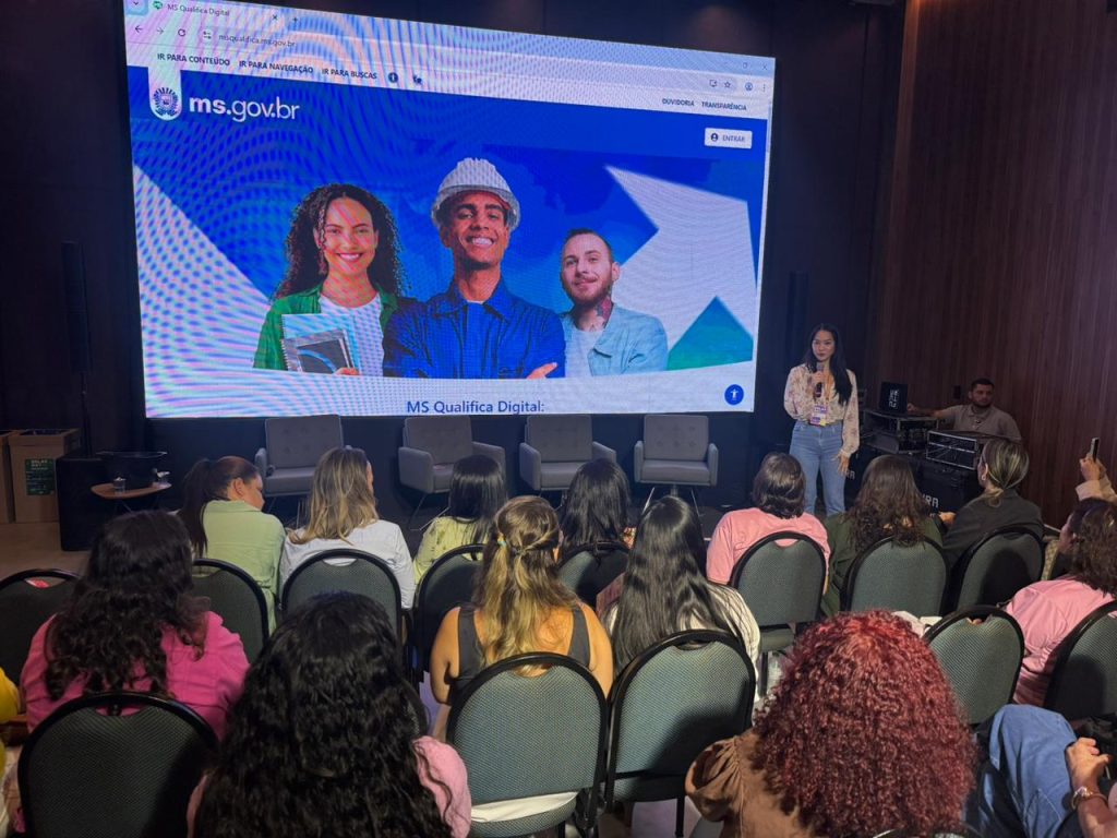 Visão da plateia de costas em direção ao palco. No centro do palco, uma mulher jovem fala ao microfone ao lado de um enorme telão de LED. O telão exibe o site "ms.gov.br" com o título "MS Qualifica Digital" e fotos de três jovens sorridentes (duas mulheres e um homem com capacete de obras). Quatro poltronas cinzas estão posicionadas vazias no palco.