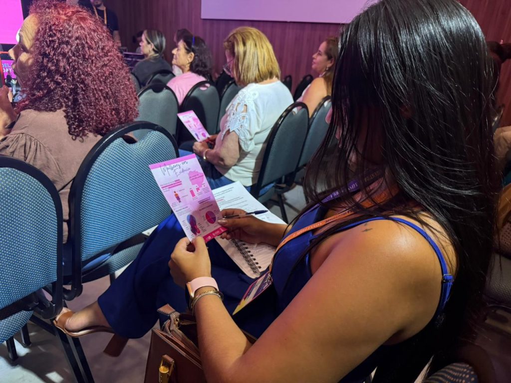 Close-up lateral de uma participante com longos cabelos pretos e blusa azul. Ela está sentada no auditório e segura um panfleto rosa intitulado "Mulheres em Evidência", que contém gráficos e informações. Ao fundo, outras mulheres aparecem desfocadas, também acompanhando a programação do evento.