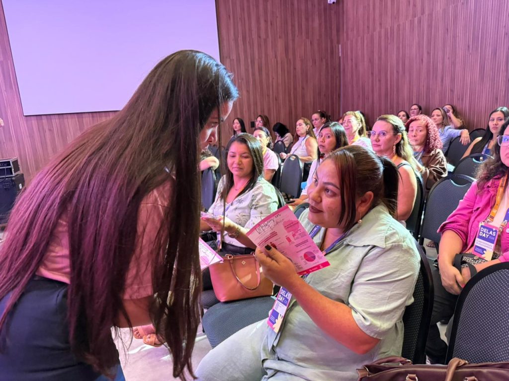Uma mulher com cabelos longos e pretos está em pé, inclinada, conversando com outra mulher sentada na plateia. A mulher sentada segura o informativo rosa do evento e olha para cima para interagir. Ambas usam crachás de identificação do "Delas Day 2026". Ao fundo, as demais participantes assistem à apresentação.