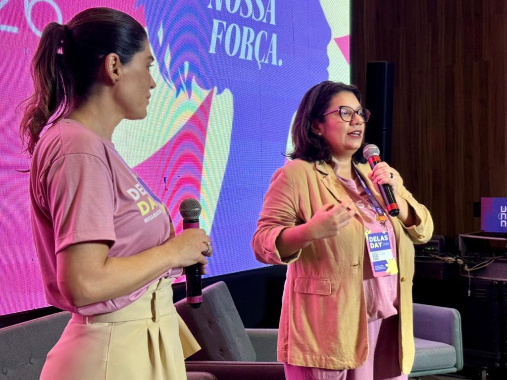 Duas mulheres no palco em frente a um telão vibrante com tons de rosa e roxo e a frase "Nossa Força". À direita, uma mulher de óculos e blazer bege fala ao microfone. À esquerda, outra mulher de camiseta rosa e saia bege segura um segundo microfone, ouvindo a colega. Elas estão em uma conversa dinâmica durante o painel.