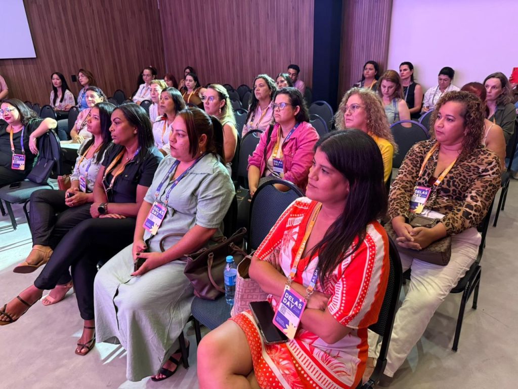 Plano médio da plateia do evento. Diversas mulheres de diferentes idades e estilos estão sentadas em fileiras, todas usando crachás azuis e amarelos do "Delas Day". A expressão de muitas é de concentração e interesse no que está sendo apresentado no palco (fora do quadro).