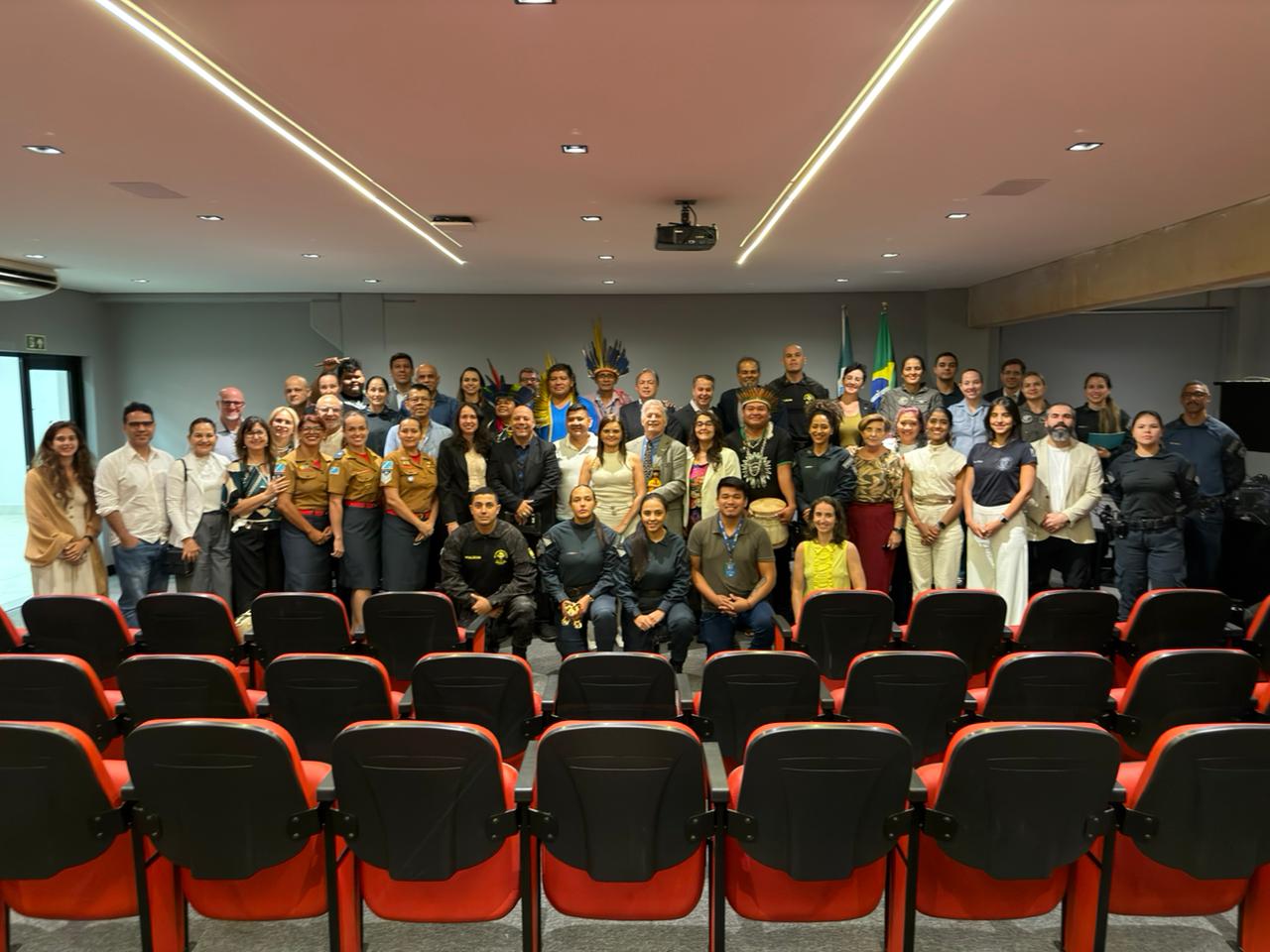 Um grande grupo de aproximadamente 50 pessoas posa para uma foto oficial em um auditório. O grupo é diverso, composto por homens e mulheres de diferentes idades. Estão presentes pessoas em trajes civis, policiais em uniformes operacionais e de gala, e lideranças indígenas com cocares tradicionais. Eles estão posicionados em pé, atrás de várias fileiras de poltronas vermelhas vazias. Ao fundo, as bandeiras do Brasil e do estado de Mato Grosso do Sul.