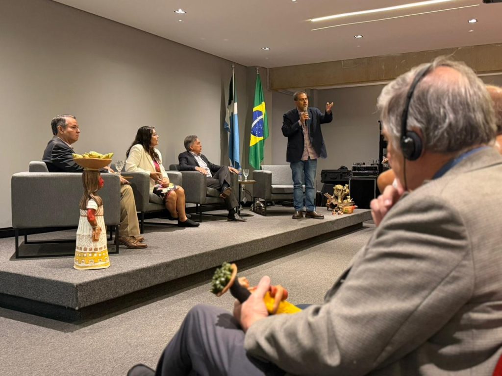 Uma visão lateral do palco durante o debate. Um homem de calça jeans e blazer escuro fala ao microfone. Os outros três integrantes do painel o observam sentados. No canto inferior direito, em primeiro plano e levemente fora de foco, vê-se um homem na plateia usando fones de ouvido para tradução simultânea, segurando uma pequena peça de artesanato indígena.