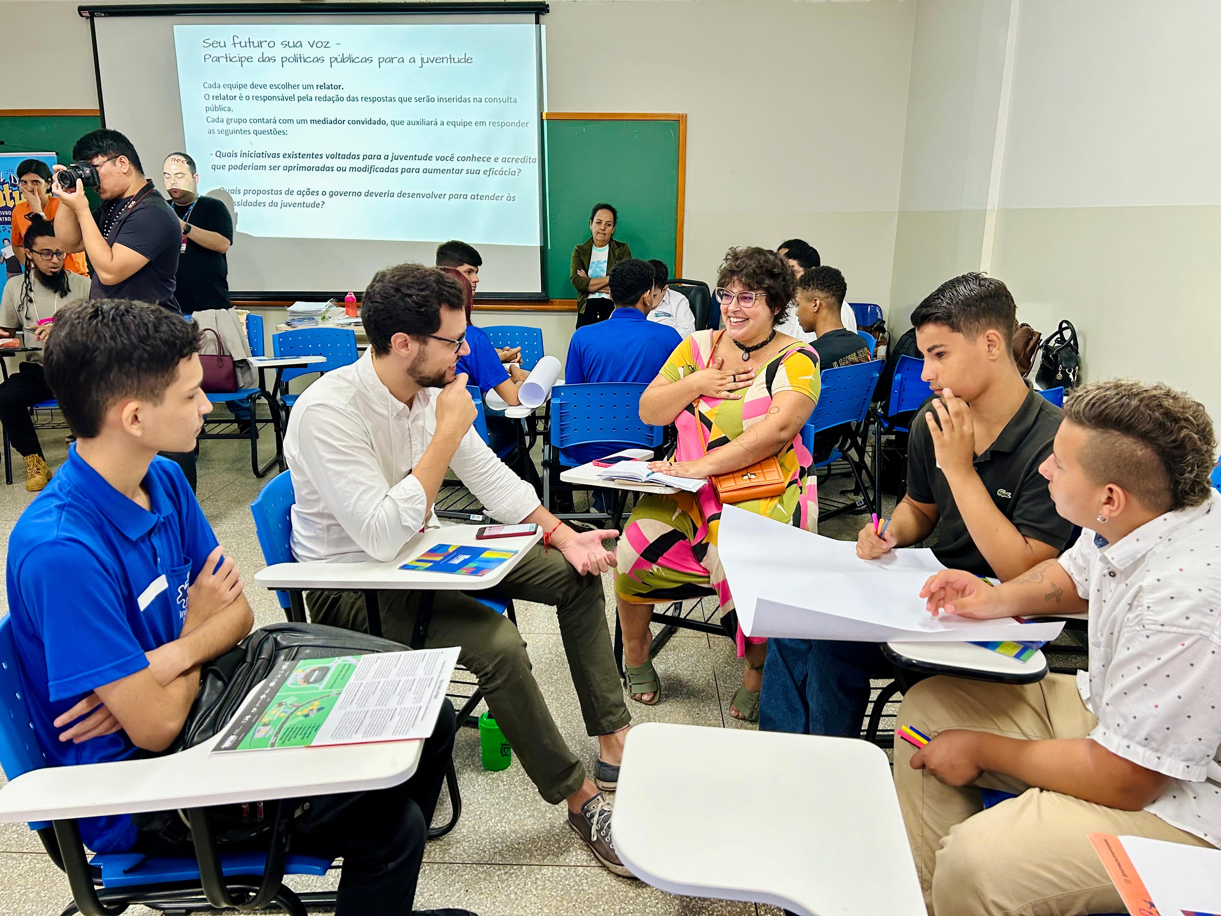 Fotografia colorida em plano médio de uma sala de aula onde jovens participam de uma oficina. Em primeiro plano, cinco pessoas estão sentadas em círculo em volta de suas classes escolares. À esquerda, um jovem de camiseta azul ouve atentamente. Ao centro, um homem de camisa branca e óculos conversa com uma mulher de vestido colorido e óculos, que sorri e gesticula com as mãos no peito. À direita, dois rapazes observam uma folha de papel em branco sobre a mesa. Ao fundo, um telão projeta o texto "Seu futuro sua voz - Participe das políticas públicas para a juventude".