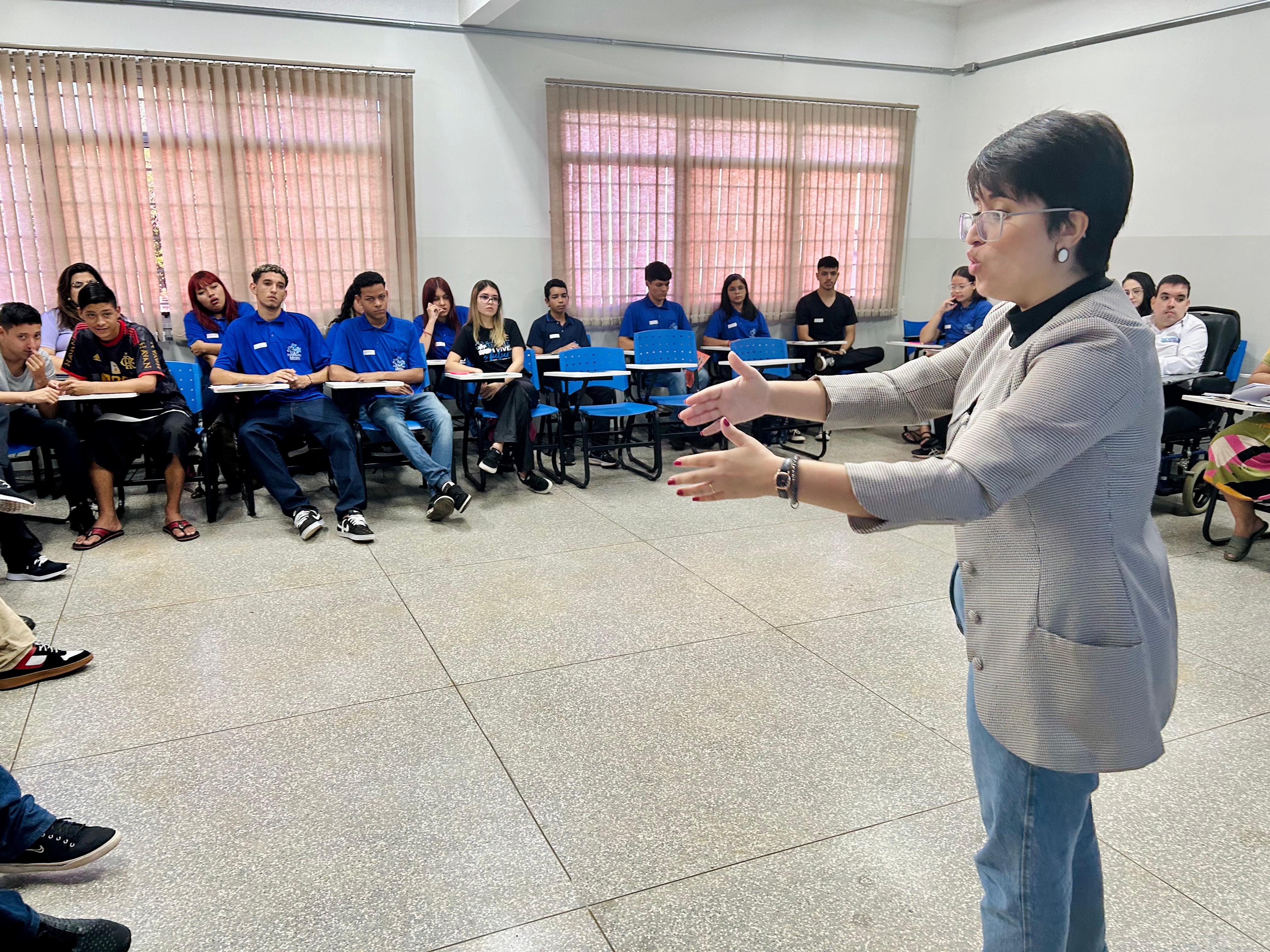 Fotografia colorida em plano aberto mostrando o interior de uma sala. À direita, em destaque e de perfil, uma mulher de cabelos curtos, óculos e blazer cinza está em pé, com os braços estendidos à frente, como se explicasse algo para o grupo. À esquerda, cerca de dez jovens estão sentados em cadeiras universitárias azuis, dispostas em semicírculo, ouvindo a apresentação. Alguns vestem camisetas azuis com um logotipo no peito. O ambiente tem paredes claras, janelas com persianas horizontais e chão de granilite claro.