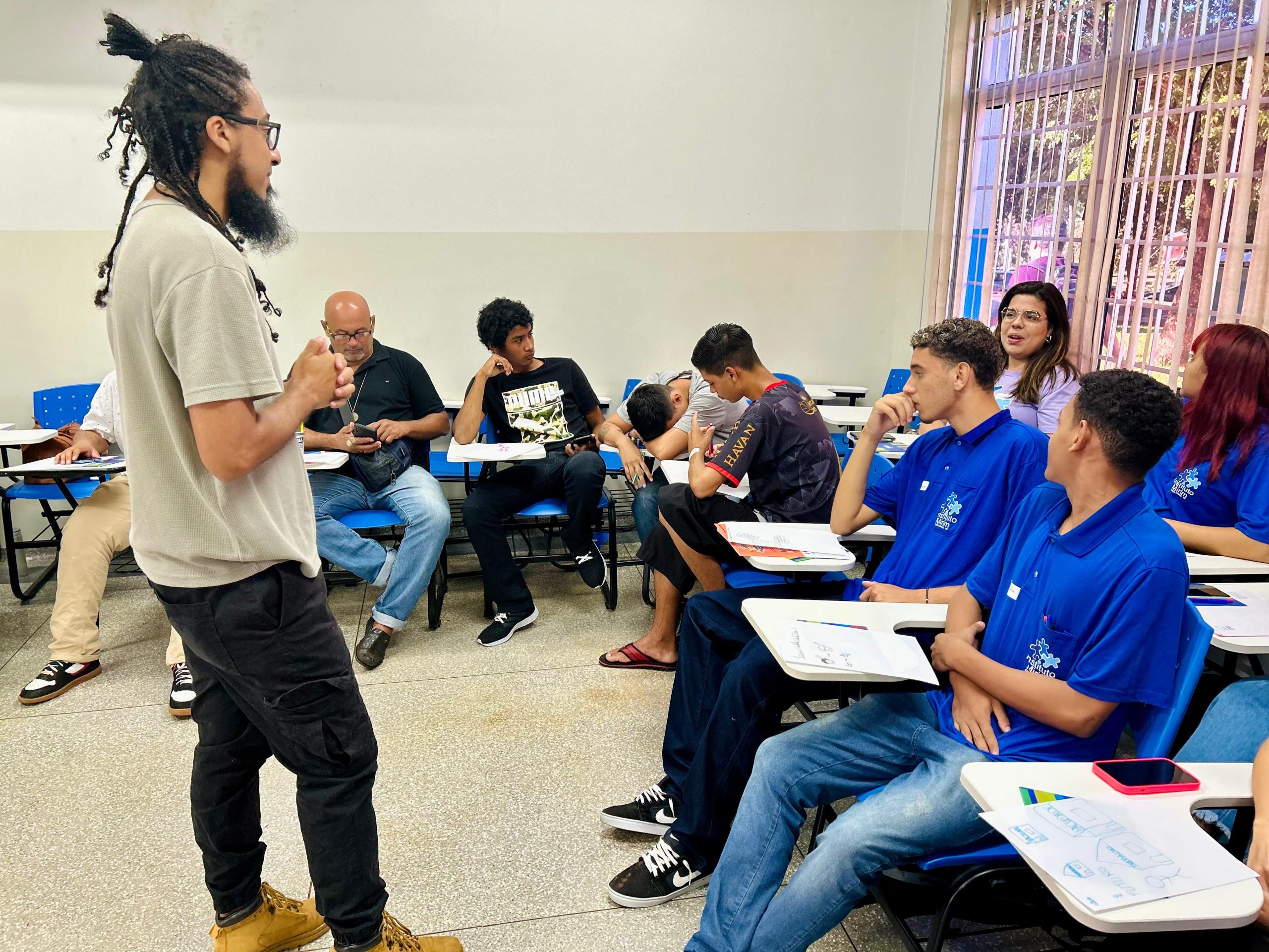 Fotografia colorida em plano médio de um momento de interação. No lado esquerdo, um homem jovem com dreadlocks presos, barba, óculos e camiseta bege está em pé, de perfil, falando com as mãos juntas. Ele se dirige a um grupo de jovens sentados em cadeiras azuis. À direita, dois rapazes de camiseta azul olham para o mediador. Entre eles, ao fundo, outras pessoas acompanham a conversa, incluindo um homem de meia-idade sentado e uma mulher de óculos que sorri. Sobre as mesas das cadeiras, há papéis com anotações e desenhos, indicando uma atividade prática em desenvolvimento.