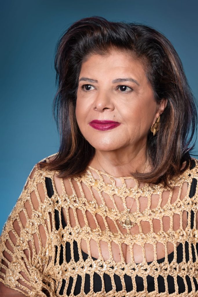 Fotografia de estúdio em plano médio de uma mulher madura, de pele clara e cabelos castanhos ondulados na altura dos ombros. Ela olha levemente para o lado, com uma expressão serena e um leve sorriso. Veste uma blusa preta sob uma sobreposição de crochê em tom dourado com tramas abertas. Ela usa batom vinho, brincos dourados em formato de flor e uma corrente fina com um pingente religioso. O fundo é de cor azul sólida.