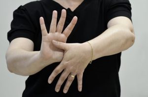 Fotografia em plano médio, do peito para baixo, de uma pessoa de pele clara vestindo uma camiseta preta. O foco principal está nas mãos da pessoa, que realiza um sinal em Libras. A mão direita está aberta, com a palma voltada para a frente e os dedos esticados e separados. A mão esquerda (que usa uma aliança dourada e uma pulseira de corrente fina) está posicionada horizontalmente à frente da outra; o polegar esquerdo toca o centro da palma da mão direita, enquanto os outros quatro dedos da mão esquerda apontam para baixo. O fundo da imagem é uma parede lisa em tom cinza claro.