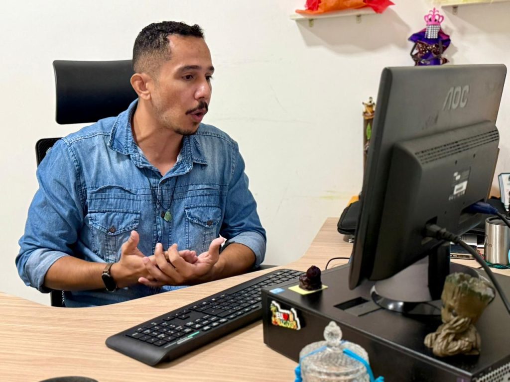 O homem aparece gesticulando com as duas mãos abertas à frente do corpo, demonstrando ênfase em sua fala durante a transmissão online. Ele continua focado na tela do computador. 