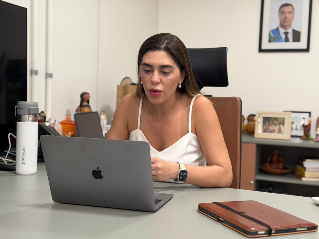 Uma mulher branca de cabelos castanhos longos e lisos, vestindo uma blusa branca de alças finas, está sentada à frente de um notebook prata. Ela olha para a tela com expressão atenta durante uma videoconferência. 