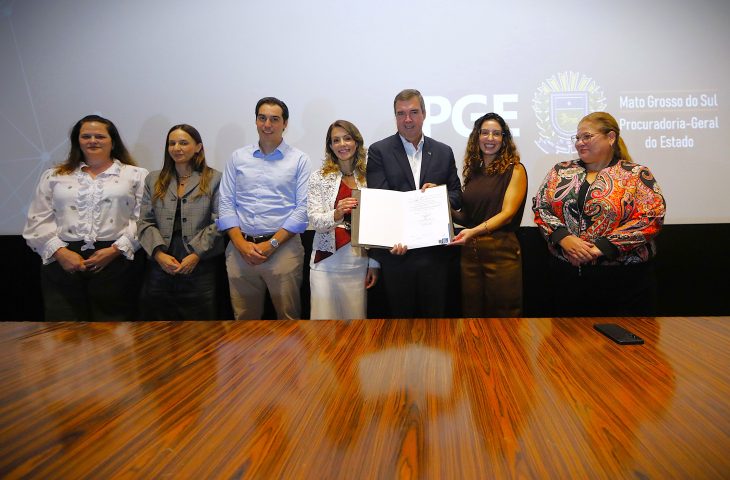 Sete pessoas estão em pé, lado a lado, em frente a um telão com o logotipo da PGE (Procuradoria-Geral do Estado de Mato Grosso do Sul). No centro, um homem de terno azul escuro e uma mulher de blusa rendada branca seguram juntos um livro de atas aberto. Todos sorriem para a câmera em um ambiente formal com uma mesa de madeira polida em primeiro plano.