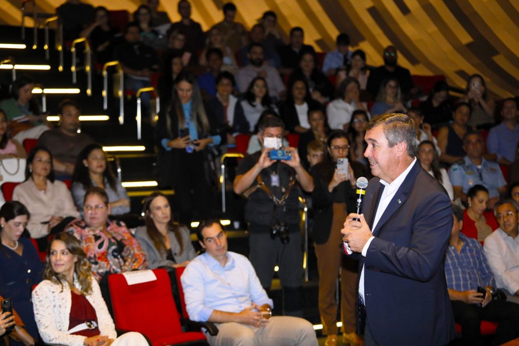 O palestrante, um homem de terno escuro, é visto de perfil falando ao microfone em primeiro plano à direita. Em foco, ao fundo, parte da plateia sentada em poltronas vermelhas o observa. Entre os espectadores, destacam-se a mulher de renda branca e um homem de camisa azul clara, ambos sentados na primeira fila.